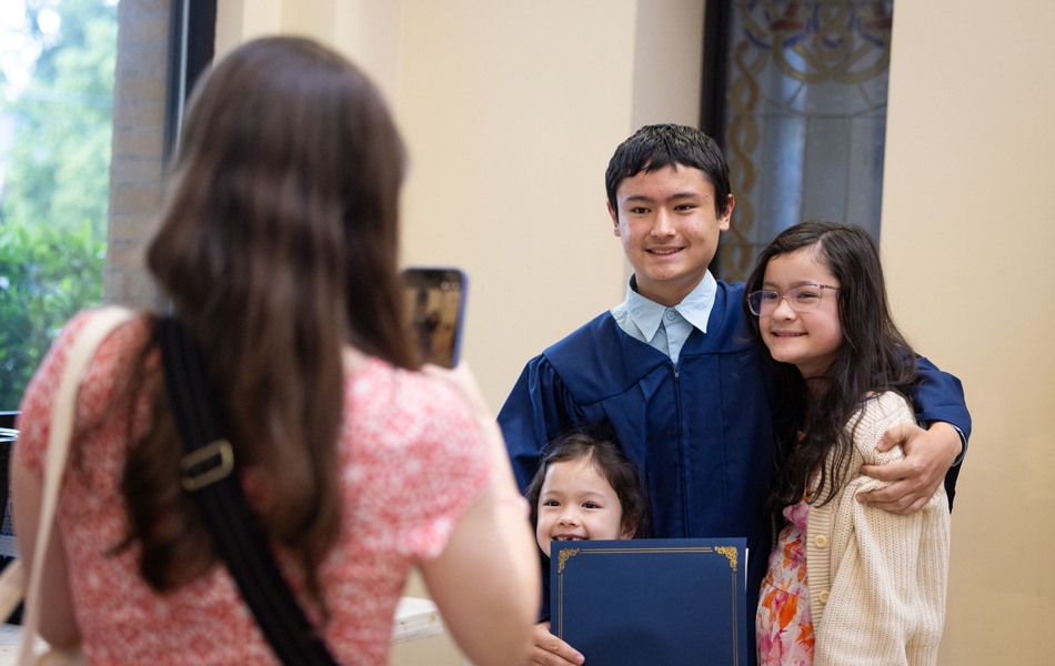 8th grade graduate in blue gown posing with his two sisters while mom takes photo on her phone