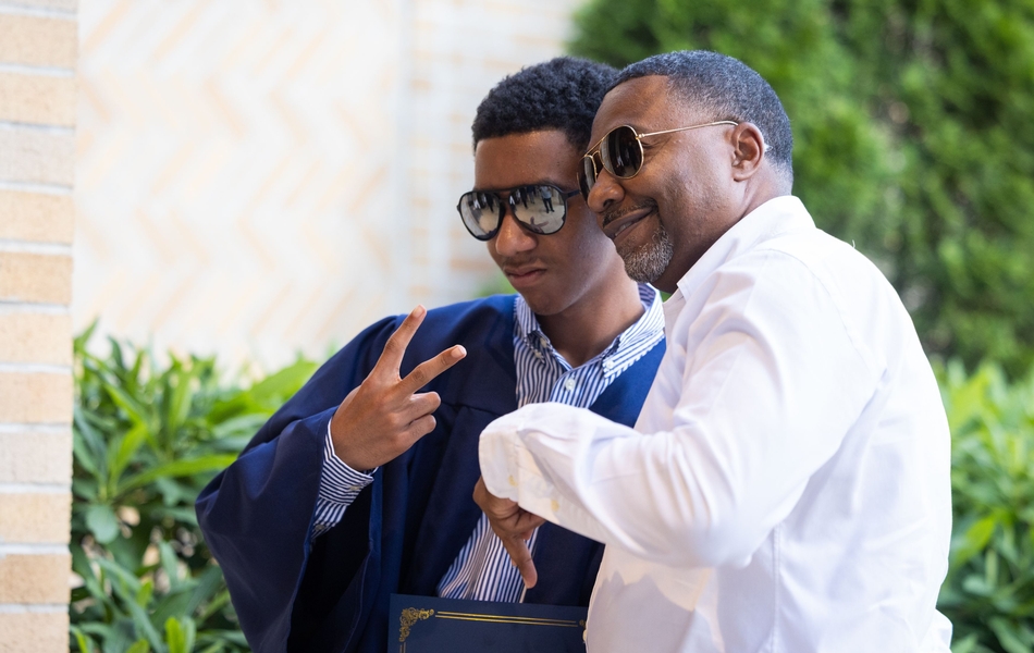 8th grade graduate in a blue gown poses with his dad while both in sunglasses and giving peace signs